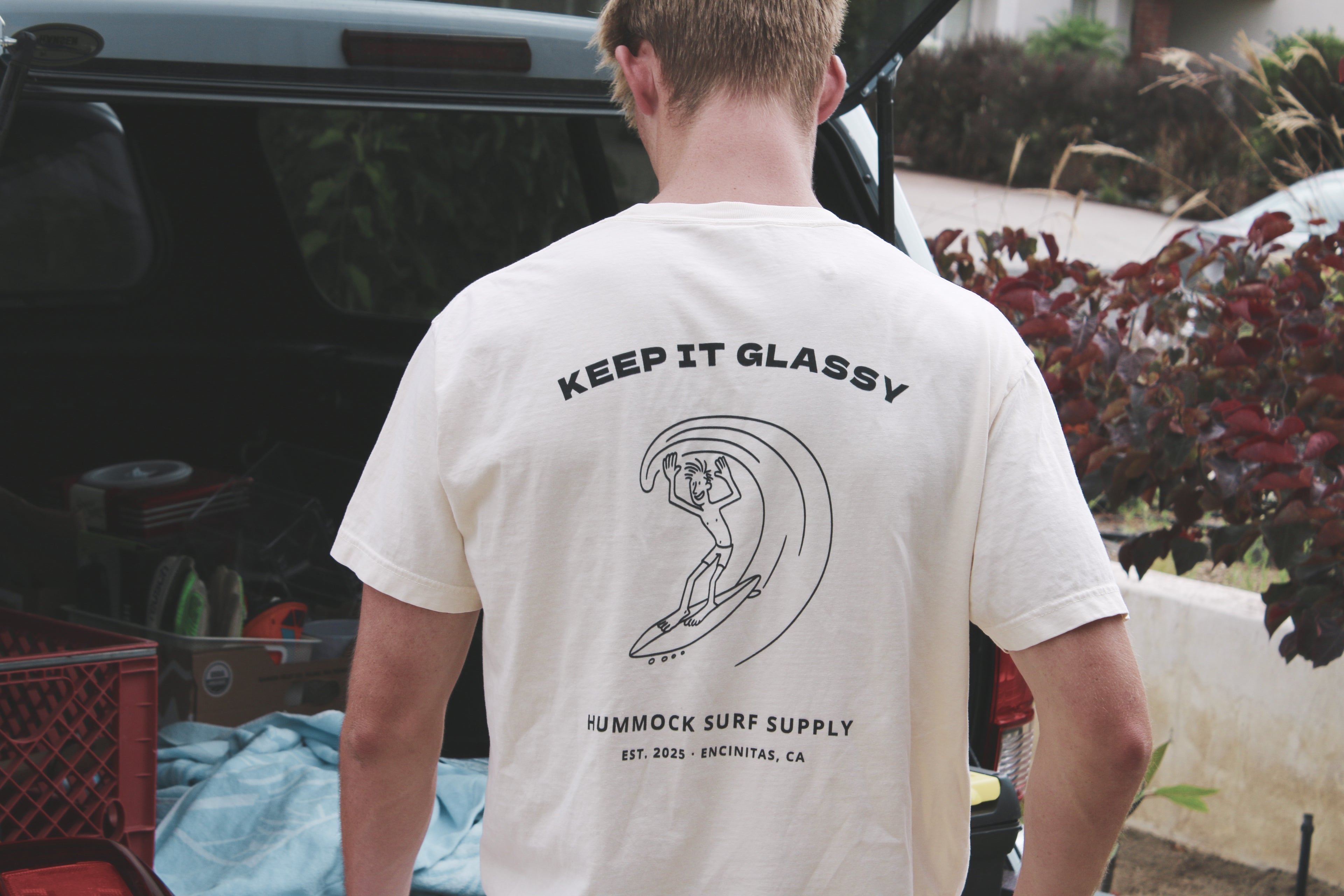Surfer wearing a t-shirt with 'Keep It Glassy' design and text, standing next to an open car trunk.